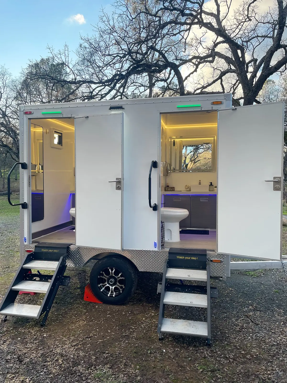Two-stall restroom trailer with both doors open, lit stalls and fold-down steps