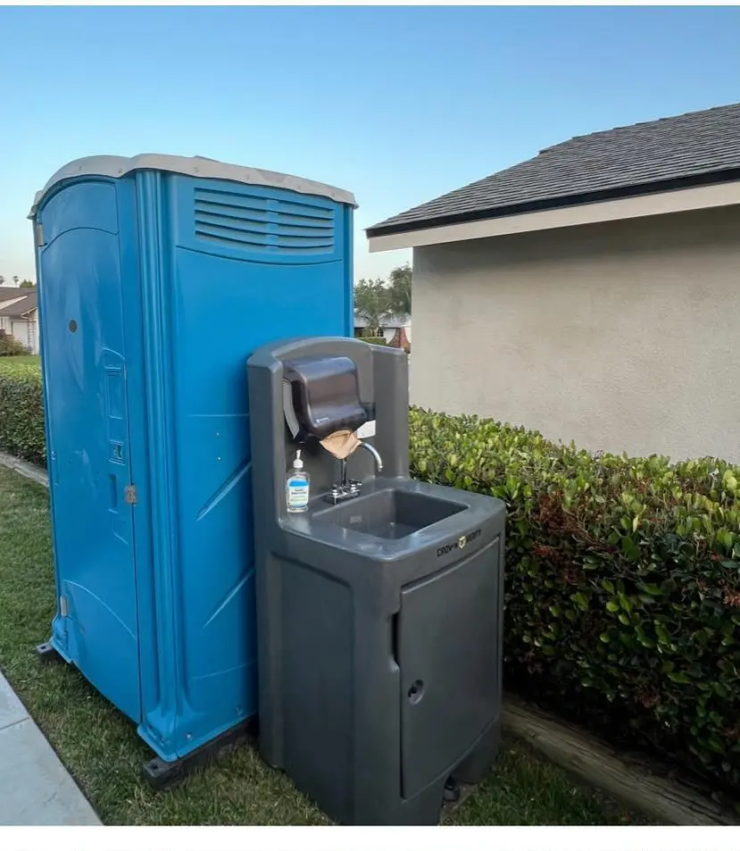 Blue porta potty with a freestanding hand washing station in a side yard